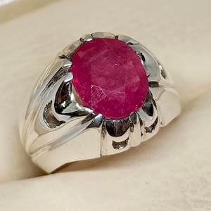 May include: A silver ring featuring a large, oval-shaped, pink ruby gemstone set in the centre. The ring has a detailed, ornate design.
