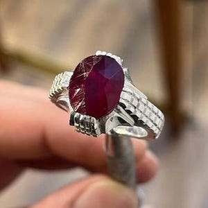 May include: A silver ring featuring a large, oval-cut ruby gemstone. The ruby is a deep red color with visible internal inclusions. The ring's band has a textured, detailed design, adding to its ornate appearance. The ring is held by a person.