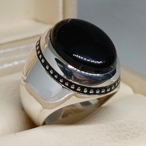Puede incluir: Un anillo de plata con una piedra ovalada negra engastada en un engaste de plata. El anillo tiene una banda decorativa con una fila de pequeñas cuentas de plata.