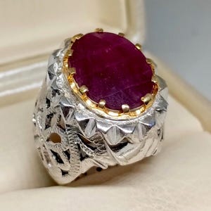 May include: A silver ring with an ornate design features a large, oval-shaped ruby gemstone set in a gold bezel. The ring has a detailed, intricate design with a filigree pattern.