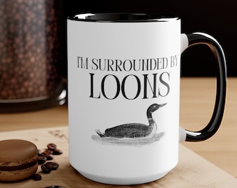 May include: White ceramic coffee mug with a black rim and handle. The mug has a black and white illustration of a loon and the text "I'm surrounded by loons".