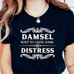 May include: A black t-shirt with white text that reads "Damsel Bout to Cause Some Distress" in a vintage-inspired font.