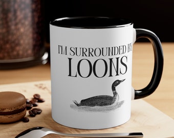 May include: White ceramic coffee mug with a black rim and handle. The mug has a black and white illustration of a loon and the text "I'm surrounded by loons".