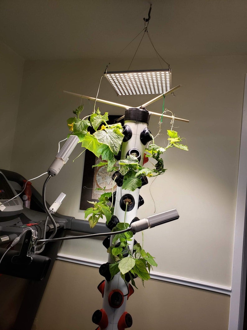Op de afbeelding: Een verticaal hydroponisch tuinsysteem met een witte toren en rode potten. De toren is omgeven door groene planten en wijnstokken. Het systeem wordt verlicht door een vierkante LED-groeilamp en twee flexibele LED-groeilampen.