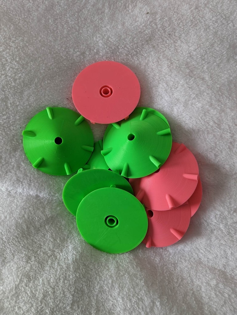 K&ouml;nnte beinhalten: Eine Ansammlung gr&uuml;ner und rosa Kunststoffscheiben. Die gr&uuml;nen Scheiben haben eine konische Form mit kleinen Vorspr&uuml;ngen am Rand. Die rosa Scheiben sind flach mit einem zentralen Loch. Die Objekte liegen auf einer wei&szlig;en, strukturierten Oberfl&auml;che.