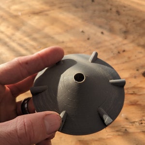 K&ouml;nnte beinhalten: Ein dunkelgraues, 3D-gedrucktes Objekt mit konischer Form und sechs gleichm&auml;&szlig;ig verteilten, dreieckigen Vorspr&uuml;ngen. Das Objekt hat ein zentrales Loch und wird in einer Hand vor einem Holzgrund gehalten.