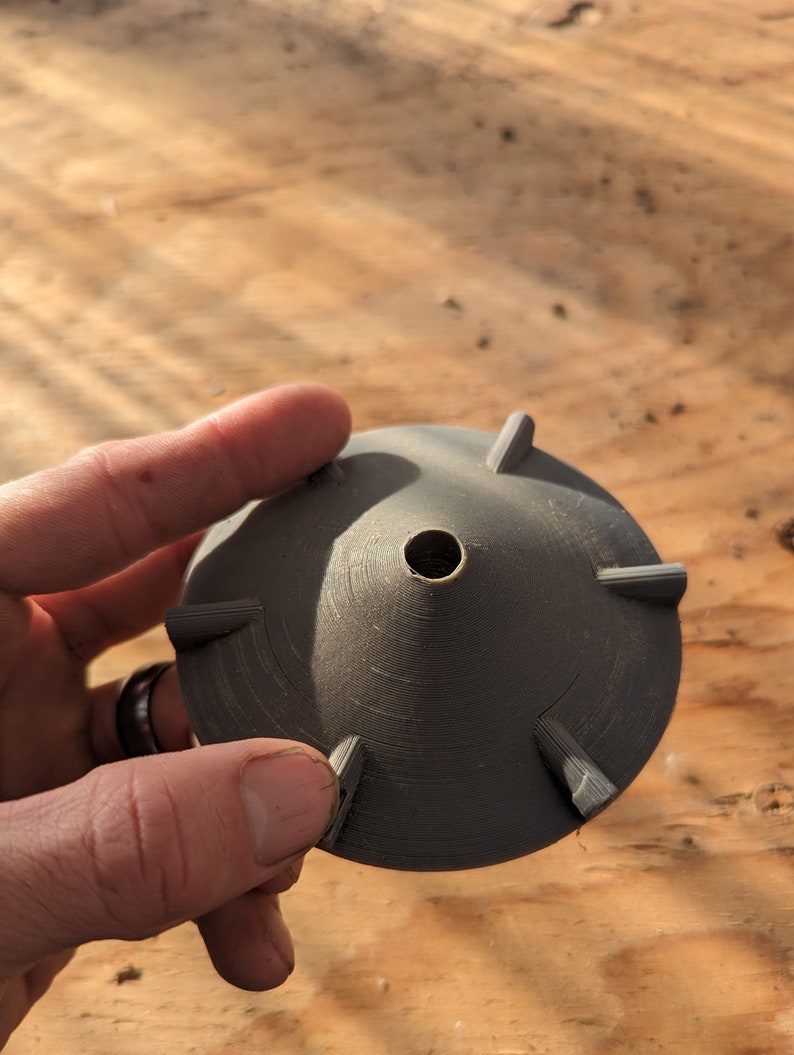 K&ouml;nnte beinhalten: Ein dunkelgraues, 3D-gedrucktes Objekt mit konischer Form und sechs gleichm&auml;&szlig;ig verteilten, dreieckigen Vorspr&uuml;ngen. Das Objekt hat ein zentrales Loch und wird in einer Hand vor einem Holzgrund gehalten.