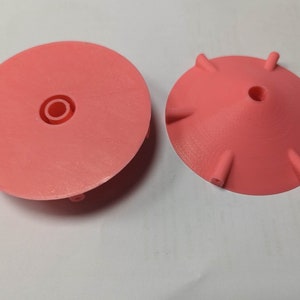 K&ouml;nnte beinhalten: Zwei korallenfarbene, 3D-gedruckte Kunststoffobjekte. Eines ist eine flache Scheibe mit einem zentralen, vertieften Kreis. Das andere ist eine konische Form mit sechs gleichm&auml;&szlig;ig verteilten, hervorstehenden Stiften am Rand und einem zentralen Loch. Beide Objekte befinden sich auf einer wei&szlig;en Oberfl&auml;che.