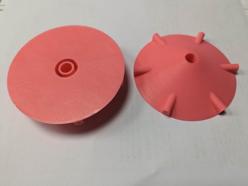 K&ouml;nnte beinhalten: Zwei korallenfarbene, 3D-gedruckte Kunststoffobjekte. Eines ist eine flache Scheibe mit einem zentralen, vertieften Kreis. Das andere ist eine konische Form mit sechs gleichm&auml;&szlig;ig verteilten, hervorstehenden Stiften am Rand und einem zentralen Loch. Beide Objekte befinden sich auf einer wei&szlig;en Oberfl&auml;che.
