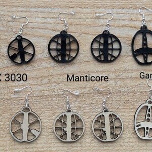Peut inclure: Un ensemble de 10 paires de boucles d'oreilles en métal noir et argent avec un motif de bobine de détecteur de métaux. Les boucles d'oreilles sont étiquetées avec les noms de différentes marques de détecteurs de métaux : Equinox, CTX 3030, Manticore, Garrett ATpro et XP Deus.