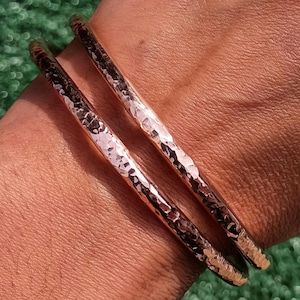May include: Two copper-colored bangles with a hammered texture are shown on a person's wrist. The bangles have a shiny, metallic finish and a slightly irregular surface. The background is a blurred green.
