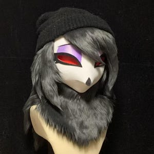 May include: A stylized creature mask featuring a smooth white face, sharp black nose, and large eyes with vibrant red irises and purple eyeshadow. The mask is adorned with thick, layered dark gray faux fur and a black knit beanie cap on top.