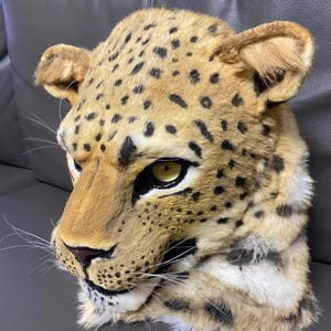 May include: A detailed, realistic leopard mask. The mask features a tan and brown spotted pattern, with a textured fur-like appearance. The eyes are a bright yellow, and the nose is a dark pink. The mask has long white whiskers and detailed ear features.