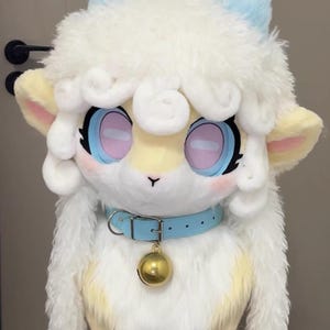 May include: A plush lamb head with large, expressive eyes and a light blue collar with a gold bell. The lamb has fluffy white fur, light blue ears, and a yellow face. The overall design is cute and cartoonish.