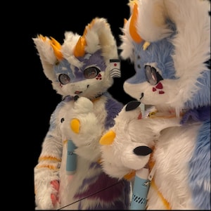 May include: A furry costume with a white, blue, purple, and yellow color scheme. The costume has large eyes, orange horns, and a detailed design. The image shows two of the costumes, with one reflected in a mirror.