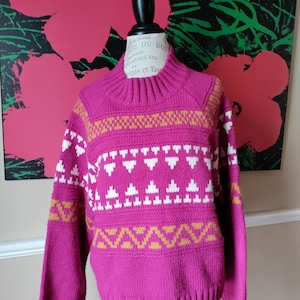 May include: A magenta sweater with a high ribbed collar and cuffs. The sweater features a geometric pattern in white and gold across the chest and sleeves. The sweater is displayed on a black mannequin.