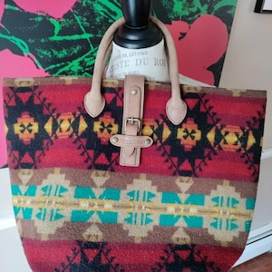 May include: A rectangular tote bag with a colorful geometric pattern in red, brown, yellow, and teal. The bag has tan leather handles and a leather strap with a buckle closure. The brand name "Pendleton" is visible on the leather strap.