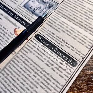 May include: A black fountain pen rests diagonally across a printed page featuring the text "WORDS TO WONDER BY" and "NOTE OF THE SEASON." The paper contains columns of text, a small illustration, and a source citation dated September 28, 2026.