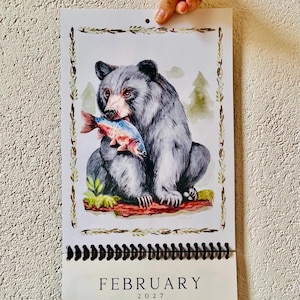 May include: A wall calendar page for February 2027 featuring a watercolor illustration of a grey bear holding a fish in its mouth. The bear sits on a log with green foliage and trees in the background, all framed by a decorative leaf border.