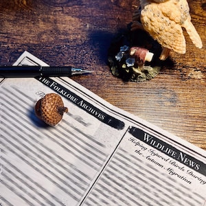 May include: A printed document titled The Folklore Archives and Wildlife News: Helping Injured Birds During the Autumn Migration, featuring a black pen, a small brown acorn, and a tan owl figurine perched on a base with small white flowers.