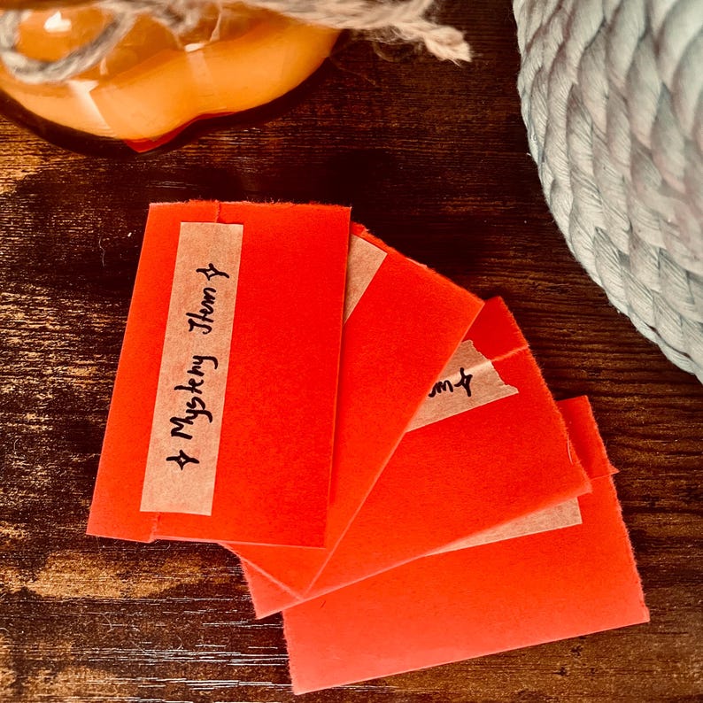 May include: A stack of four rectangular bright orange paper envelopes. The top envelope features a strip of light brown adhesive tape with the handwritten text Mystery Item and two small star symbols drawn in black ink.