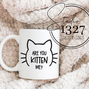 May include: White ceramic mug with a black outline of a cat face and the text "Are you kitten me?" printed on it.