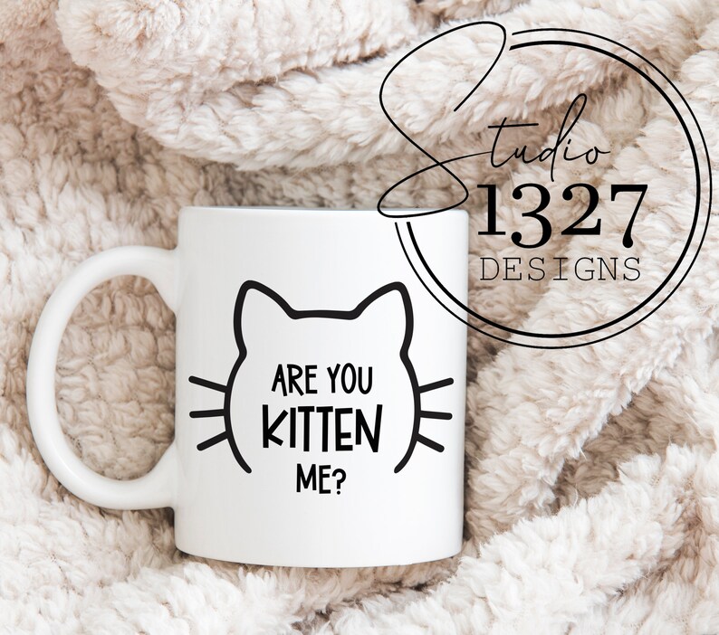 May include: White ceramic mug with a black outline of a cat face and the text "Are you kitten me?" printed on it.