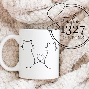 May include: White ceramic mug with a black line drawing of two cats with their tails intertwined forming a heart shape. The mug is on a white fuzzy surface.