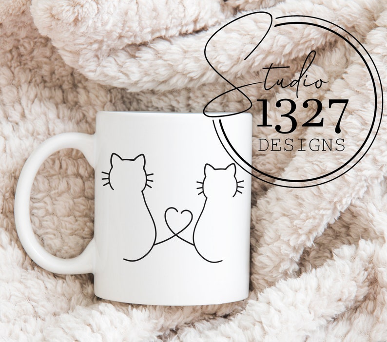 May include: White ceramic mug with a black line drawing of two cats with their tails intertwined forming a heart shape. The mug is on a white fuzzy surface.