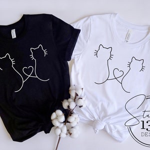 May include: Two black and white t-shirts with a simple line drawing of two cats with their tails intertwined to form a heart. The shirts are laid out on a white surface with a sprig of cotton.