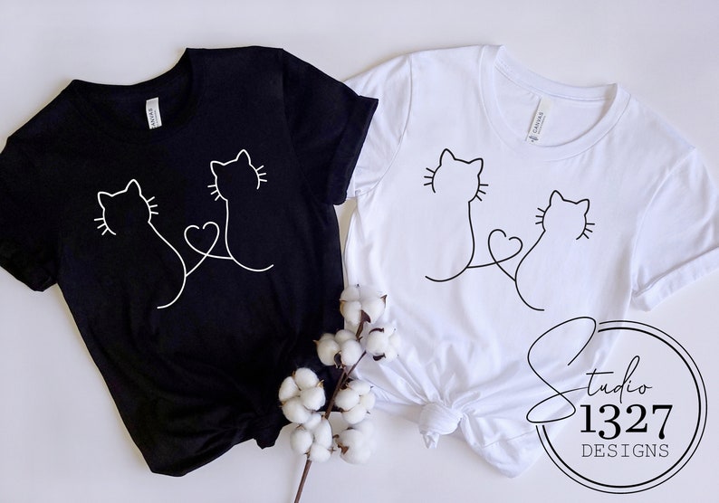 May include: Two black and white t-shirts with a simple line drawing of two cats with their tails intertwined to form a heart. The shirts are laid out on a white surface with a sprig of cotton.