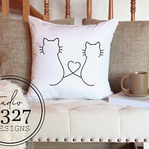 May include: White throw pillow with a black line drawing of two cats with their tails intertwined to form a heart shape.
