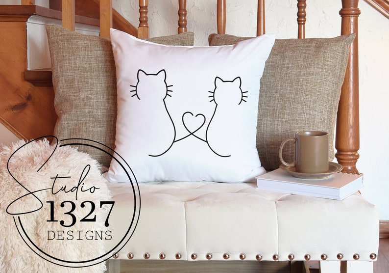 May include: White throw pillow with a black line drawing of two cats with their tails intertwined to form a heart shape.