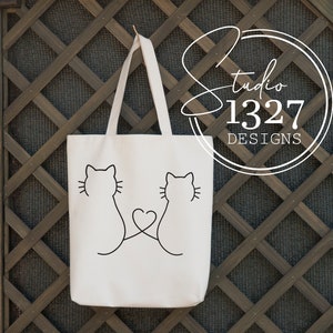 May include: White tote bag with black outline of two cats facing each other with a heart in the middle. The text "Studio 1327 Designs" is on a circle in the upper right corner.