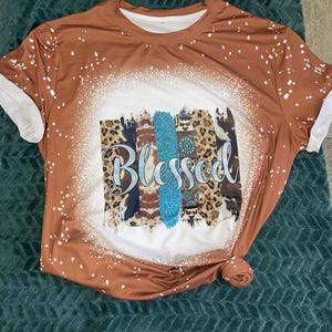 Könnte beinhalten: Ein braunes T-Shirt mit weißen Ärmeln und einem gebleichten Design. Das Shirt zeigt das Wort "Blessed" in hellblauer Schrift über einem Design mit Leopardenmuster, Türkis und marineblauen Streifen.
