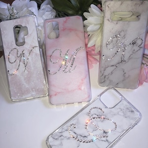 May include: Four phone cases with marble designs and rhinestone initial letters. The cases feature the letters "W", "M", "K", and "S". The marble patterns are in shades of pink, white, and grey.