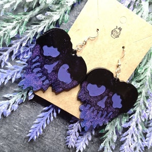Könnte beinhalten: Ein Paar schwarze und lila Glitzer-Schädel-Ohrringe mit silbernen Haken. Die Ohrringe befinden sich auf einer braunen Karte mit einem kleinen Logo.