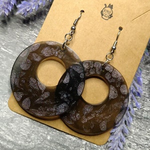 Könnte beinhalten: Ein Paar silberne Creolen mit einem schwarz-weißen Muster. Die Ohrringe sind aus Harz und haben eine glänzende Oberfläche.