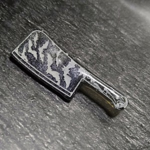 May include: A silver-colored cleaver with a black handle and a white design on the blade.