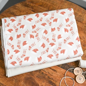 Könnte beinhalten: Ein weißer Stoff mit einem roten Muster aus Strümpfen und ein weißer Stoff mit einer einfachen Webart. Es gibt eine Spule weißen Fadens und drei Holzknöpfe auf einer Holzoberfläche.