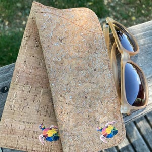 Peut inclure: Un étui à lunettes de soleil en liège avec deux motifs de tortues colorées et une paire de lunettes de soleil marron clair. L'étui est en liège texturé marron clair. Les lunettes ont une monture marron clair et des verres foncés.