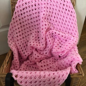 May include: A pink crochet blanket, with an open weave design, is placed inside a wicker basket. The basket has a rectangular shape and black leather straps. The blanket is folded, creating a soft, textured appearance. The basket is made of woven material.