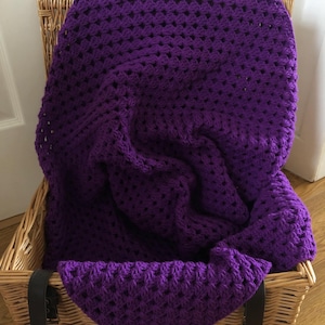 May include: A deep purple crochet blanket featuring a repeating open-work stitch pattern. The textile is draped inside a light brown woven wicker basket that has two dark brown leather straps attached to the front rim of the container.