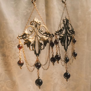 May include: A pair of gold-toned earrings featuring ornate, filigree-style metal leaf components suspended from lever-back hooks. Each earring is adorned with multiple delicate gold chains and dark red, spherical gemstone beads.