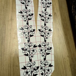 Könnte beinhalten: Zwei florale Zweigaufkleber mit schwarzen Blüten und Blättern. Die Aufkleber befinden sich auf einem weißen Hintergrund mit einem roten Gittermuster. Die Aufkleber sind aus einem Materialbogen geschnitten.