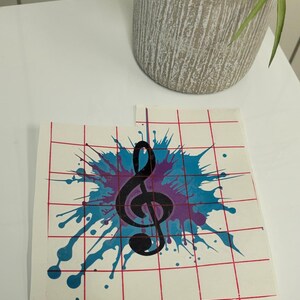 May include: A black treble clef graphic on a white background with a blue and purple paint splatter design. The graphic is on a sheet of transfer paper with a grid pattern.