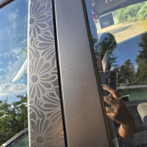 K&ouml;nnte beinhalten: Nahaufnahme eines Autot&uuml;rrahmens mit einem dekorativen Blumenmuster. Der Rahmen ist dunkelgrau, und das Blumendesign ist in die Oberfl&auml;che ge&auml;tzt. Das Fenster spiegelt die Umgebung wider.