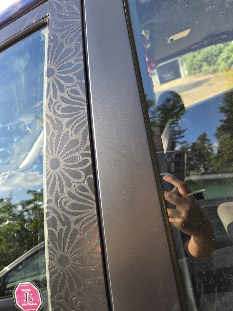 K&ouml;nnte beinhalten: Nahaufnahme eines Autot&uuml;rrahmens mit einem dekorativen Blumenmuster. Der Rahmen ist dunkelgrau, und das Blumendesign ist in die Oberfl&auml;che ge&auml;tzt. Das Fenster spiegelt die Umgebung wider.