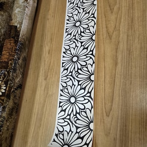 K&ouml;nnte beinhalten: Ein langer, wei&szlig;er Aufkleber mit einem sich wiederholenden schwarzen Blumenmuster. Die Blumen haben abgerundete Bl&uuml;tenbl&auml;tter und sind vertikal angeordnet. Der Aufkleber ist teilweise abgezogen und zeigt eine Holzoberfl&auml;che.