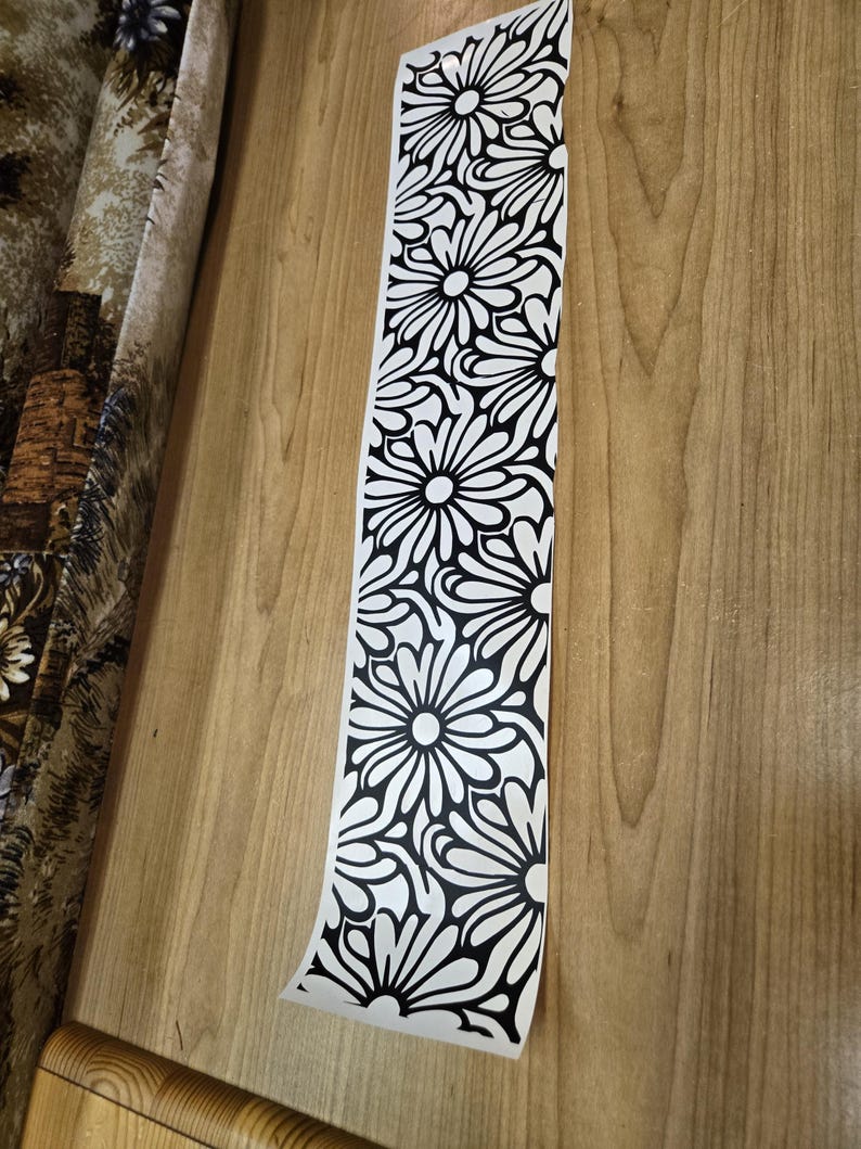 K&ouml;nnte beinhalten: Ein langer, wei&szlig;er Aufkleber mit einem sich wiederholenden schwarzen Blumenmuster. Die Blumen haben abgerundete Bl&uuml;tenbl&auml;tter und sind vertikal angeordnet. Der Aufkleber ist teilweise abgezogen und zeigt eine Holzoberfl&auml;che.